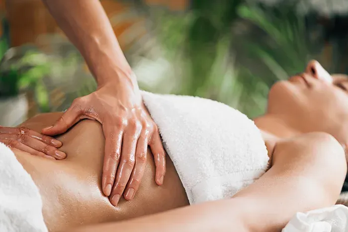 Main huilée posée sur le ventre d’une personne allongée, recouverte de serviettes blanches, pendant un massage abdominal